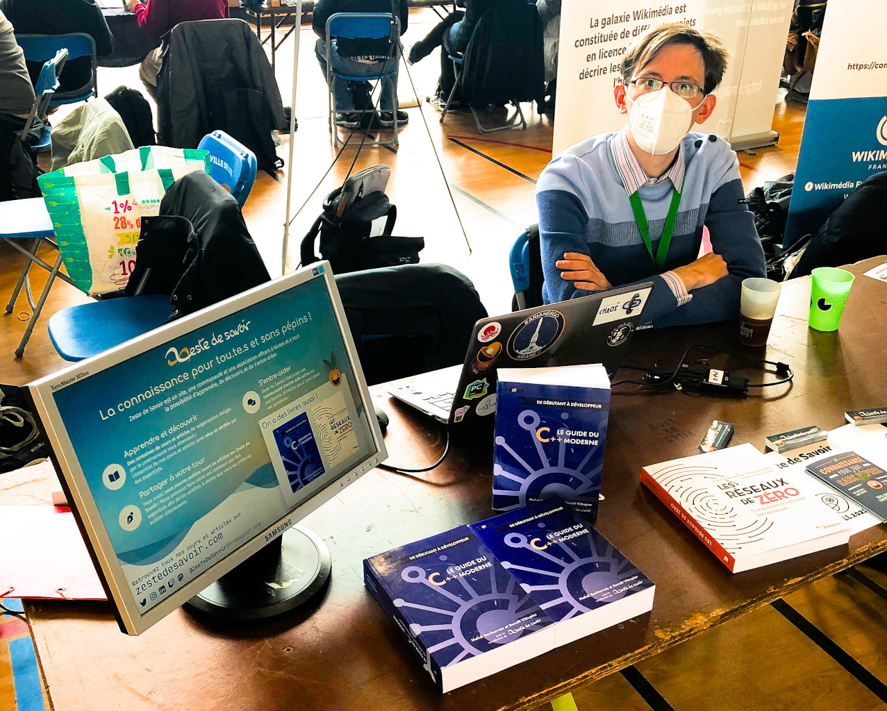 Photo du stand de Zeste de Savoir aux Journées du Logiciel Libre de Lyon. On y voit une affiche numérique expliquant ce qu'est Zeste de Savoir (apprendre, découvrir, s'entre-aider, et partager à son tour) ainsi que nos réseaux (site web, réseaux sociaux) et livres (« Le guide du C++ moderne » et « Les réseaux de Zéro »). À côté, sont éparpillés sur le stand des exemplaires des deux livres, des flyers et des autocollants, et deux tasses de thé.